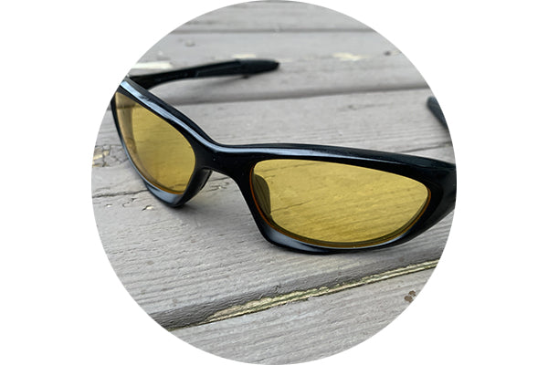 A close-up view of tracer yellow lenses in sunglasses that are laying on a wooded surface.