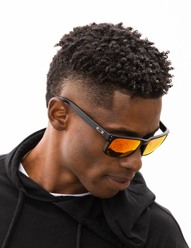 Shoulders-up view of a model with short, dark hair looking down and wearing sunglasses with Fire red lenses
